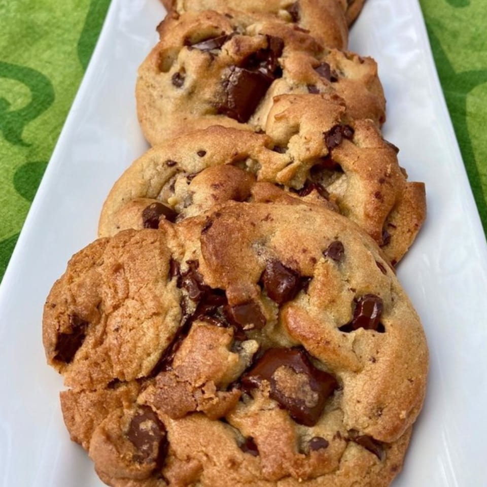 Four warm, golden-brown chocolate chip cookies with melted chocolate chunks on a white rectangular plate.