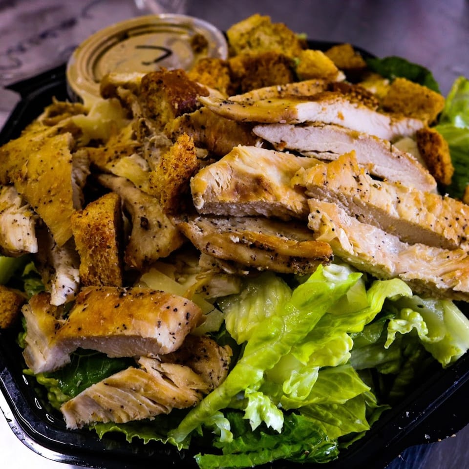 Capitini's Italian Deli's Caesar salad with grilled chicken, croutons, and dressing in a black container.