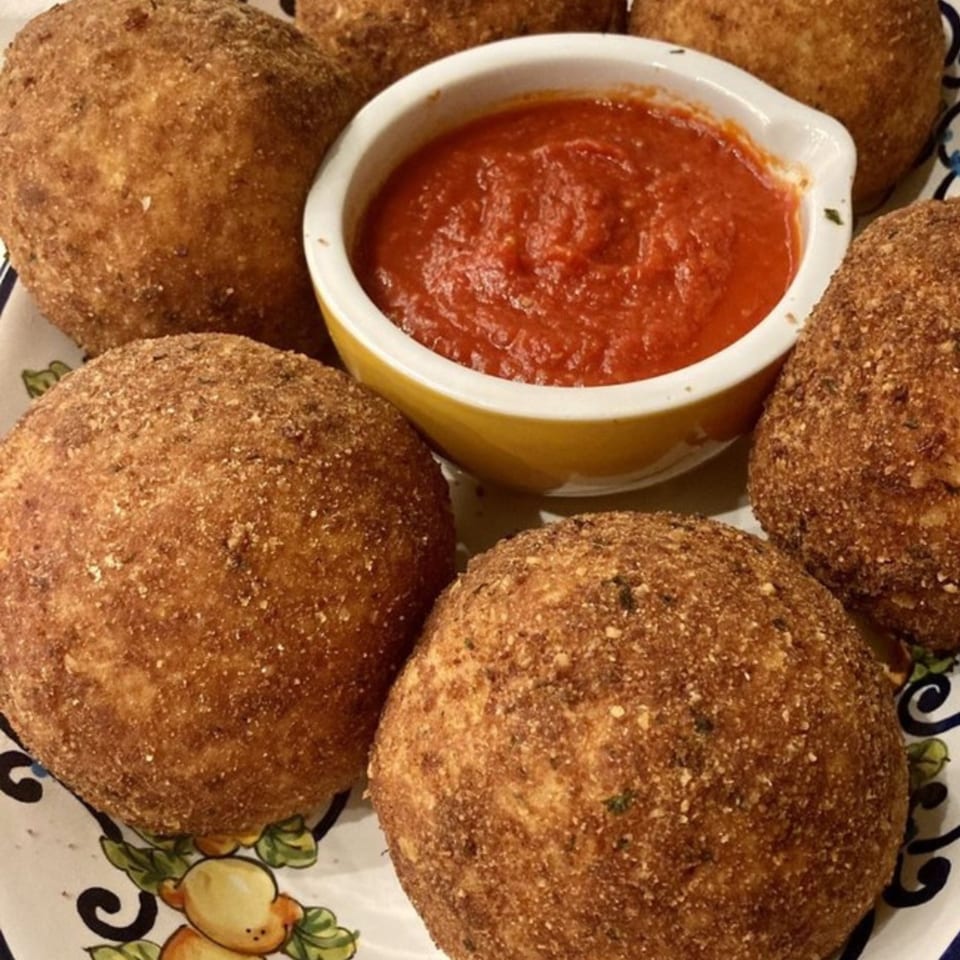 Golden-brown arancini, Italian fried rice balls, surround a bowl of red marinara sauce on a decorative plate.